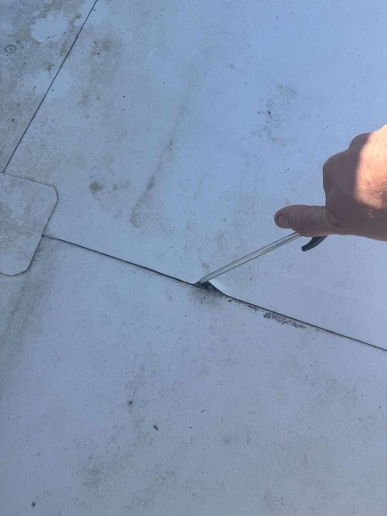 A hand lifting an open lap seam on a white flat-roof membrane with a probe